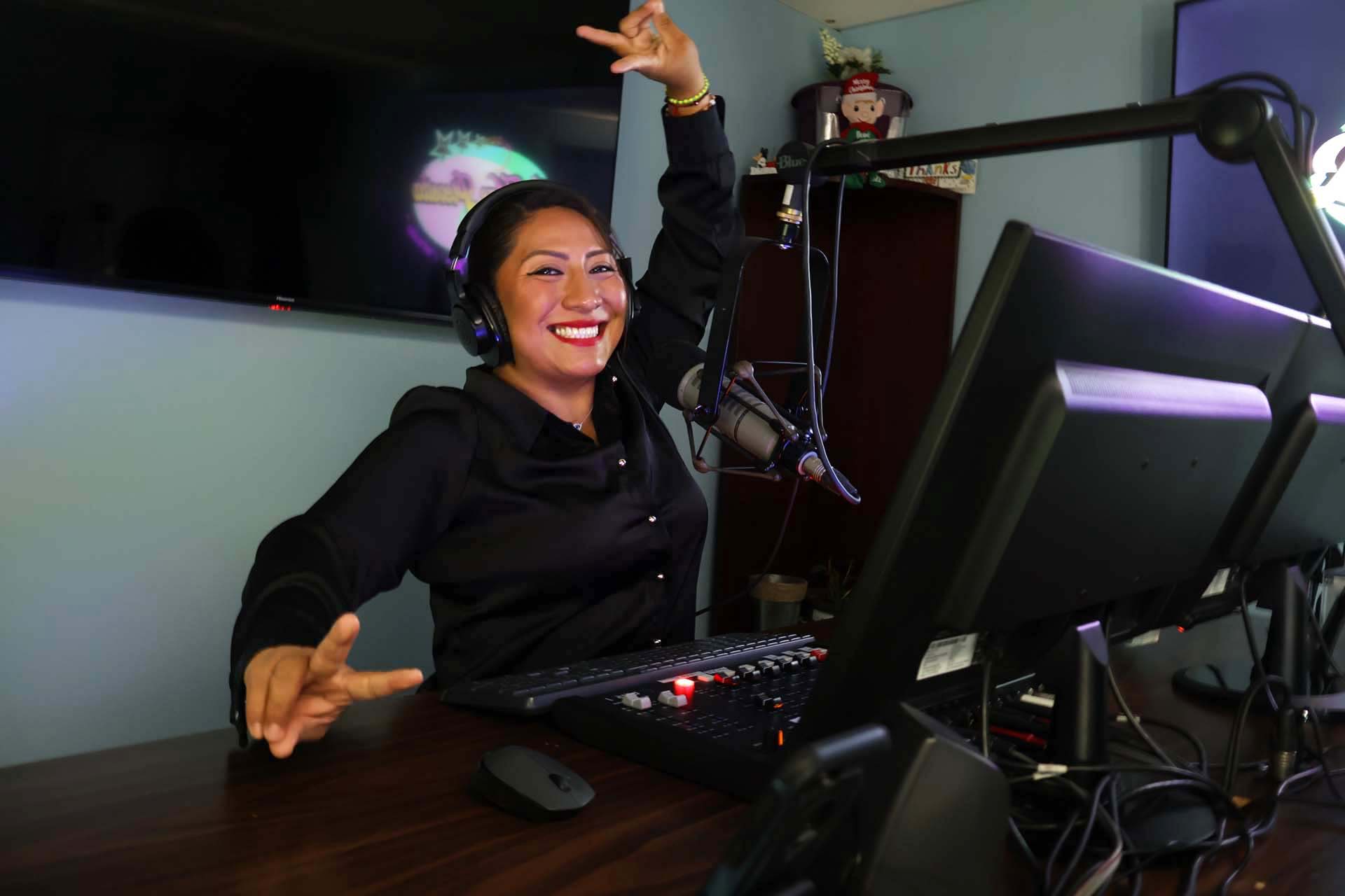 Monica Acuña en cabina bien sonriente conectando con sus oyentes que escuchan la radio por el celular.